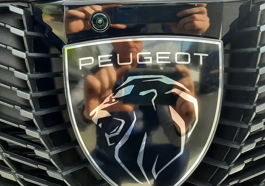 Gros plan sur le logo Peugeot noir et argent sur la calandre noire du 3008 gris titane, avec reflet visible.