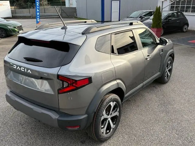 Vue 3/4 arrière droite du Dacia Duster 1.8 Hybrid 155 Journey en gris schiste métallisé avec jantes bi-ton et barres de toit.