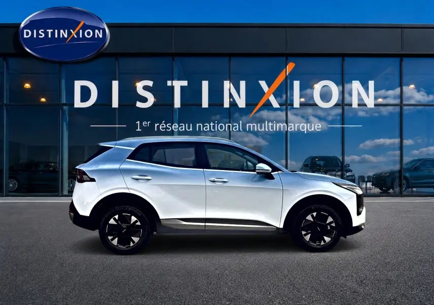 Profil droit d’un KIA SPORTAGE blanc 2026 hybride, avec jantes noires et toit légèrement incliné, devant une vitrine Distinxion.