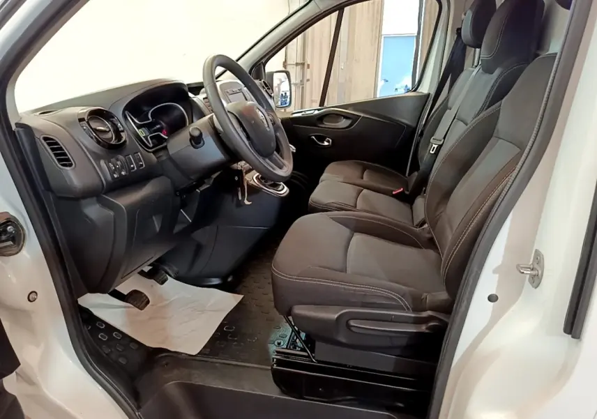Vue intérieure côté conducteur du Renault Trafic Fourgon blanc, montrant les sièges avant et le tableau de bord noir.