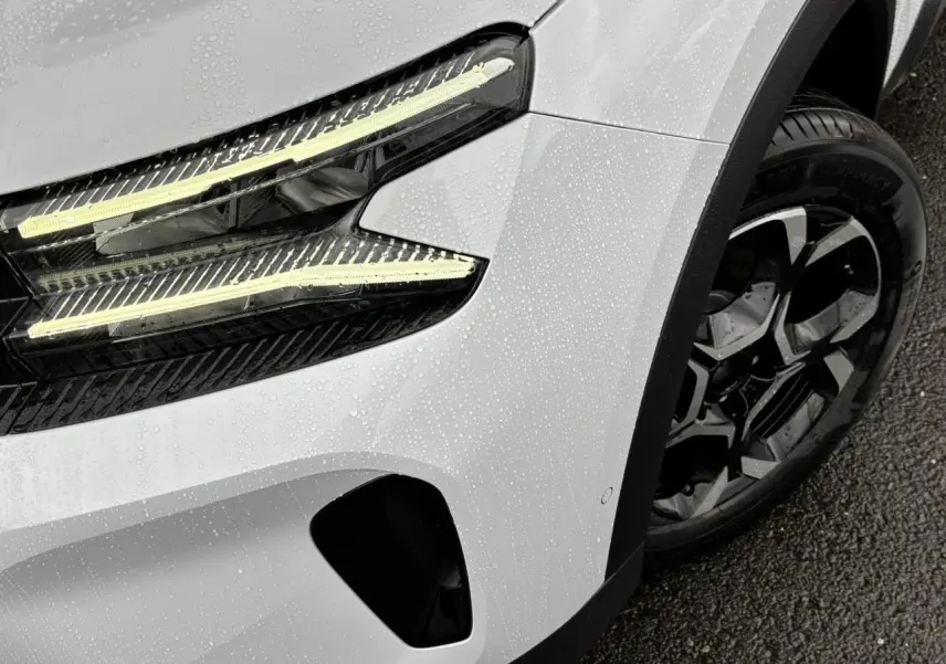 Gros plan sur le phare avant gauche allumé et la jante alliage noire du Citroën C5 Aircross blanc Okénite sous la pluie.
