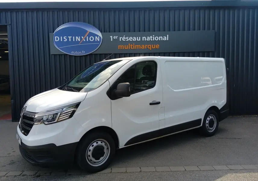 Renault Trafic Fourgon blanc en 3/4 avant droit, avec calandre noire et jantes acier, stationné devant un bâtiment gris.