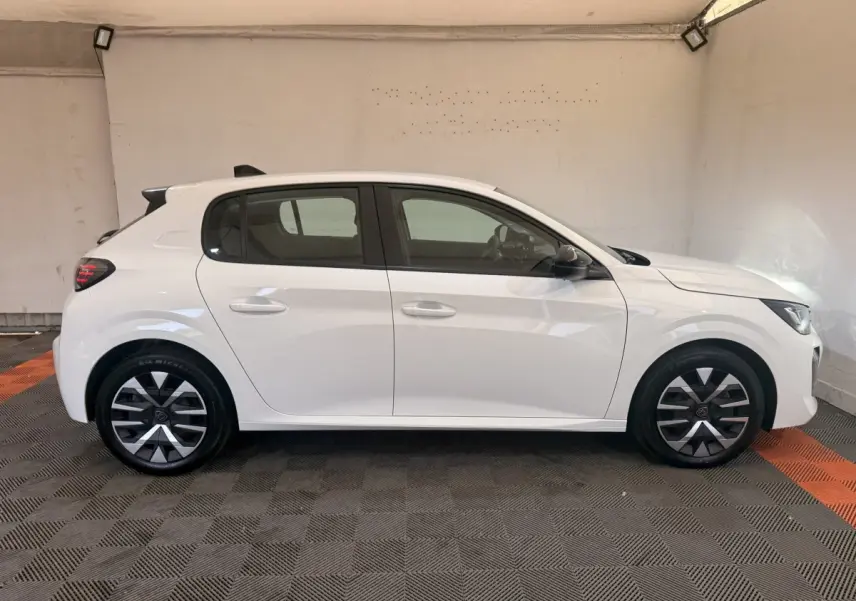 Profil droit d'une Peugeot 208 blanche 2025 avec jantes alliage et vitres teintées dans un garage.