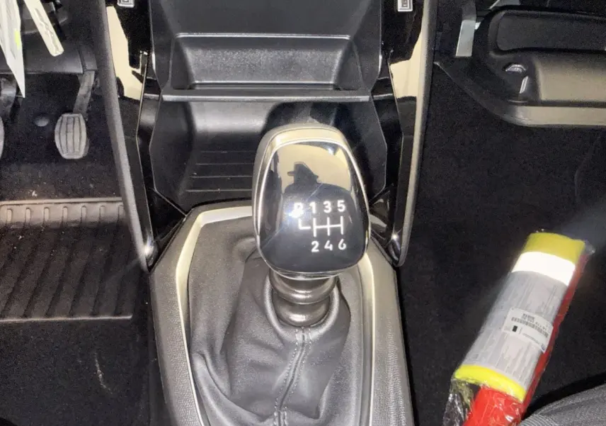 Gros plan sur le levier de vitesse manuel noir dans l’habitacle d’une Peugeot 208 blanche, avec pédales et extincteur visibles.