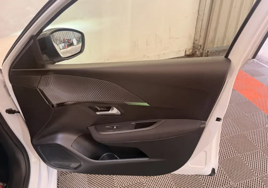 Vue côté gauche de la porte avant blanche d'une Peugeot 208 2025, avec garniture noire et insert effet carbone.