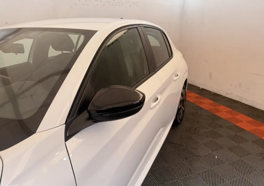 Vue côté gauche arrière d'une Peugeot 208 blanche avec rétroviseur noir et jantes sportives visibles.