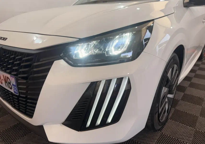Gros plan sur l'avant droit d'une Peugeot 208 blanche 2025 avec phares LED allumés et signature lumineuse verticale.