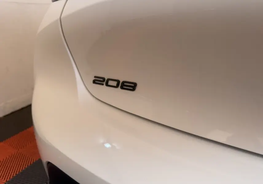 Gros plan sur le logo 208 à l'arrière gauche d'une Peugeot 208 blanche, avec sol en damier noir et orange.