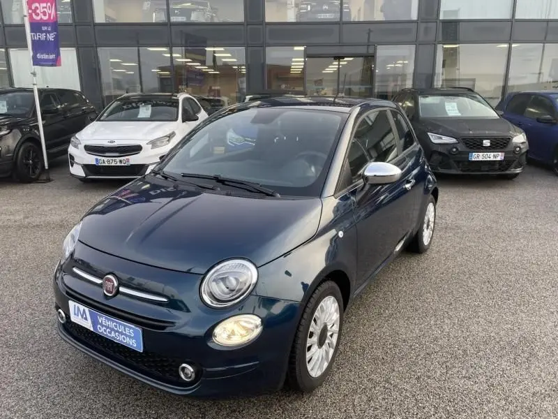 Vue 3/4 avant droite d'une FIAT 500 bleu métal 2023 avec jantes argentées et rétroviseurs chromés, stationnée devant un showroom.