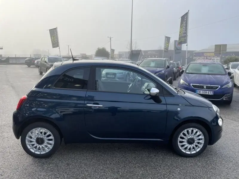 Vue de profil côté gauche d'une Fiat 500 2023 bleu métal avec jantes alliage et rétroviseurs blancs.