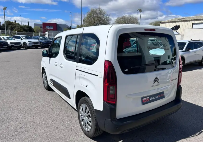 Vue 3/4 arrière droite d'un Citroën Berlingo blanc avec barres de toit et vitres teintées, stationné en extérieur.