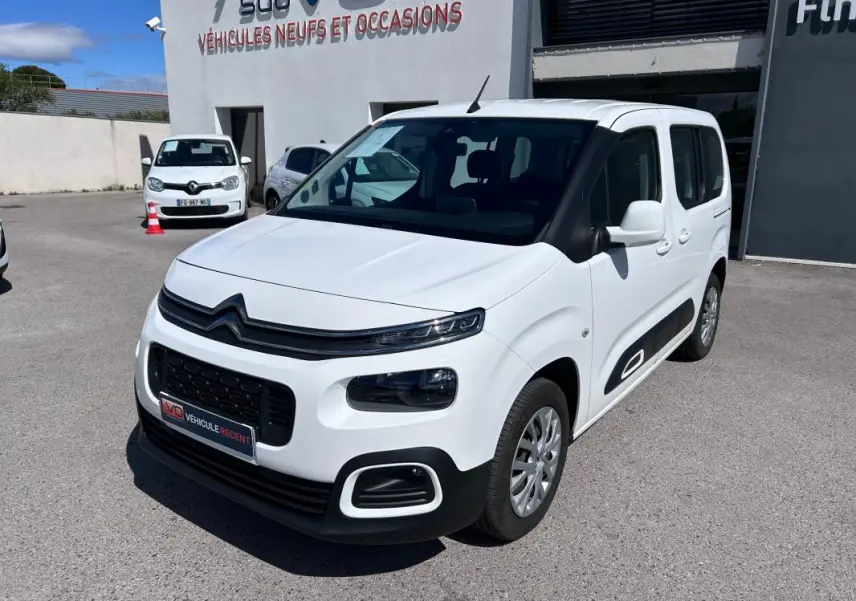 Citroën Berlingo blanc en 3/4 avant droit, avec barres de toit et protections latérales noires visibles.