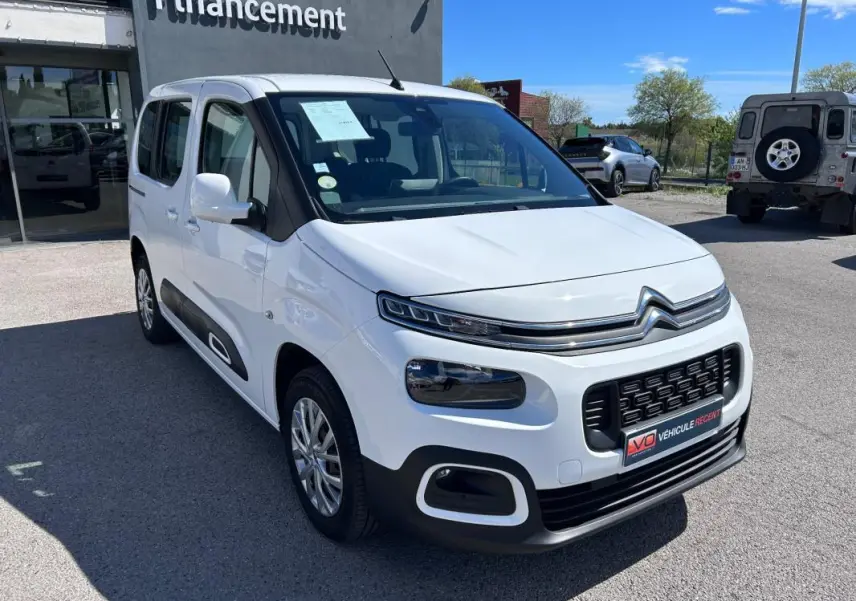 Vue 3/4 avant droite d'un Citroën Berlingo blanc avec barres de toit et protections latérales noires.