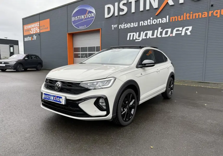 Volkswagen Taigo blanc en 3/4 avant droit avec jantes noires, stationné devant un bâtiment gris et orange.