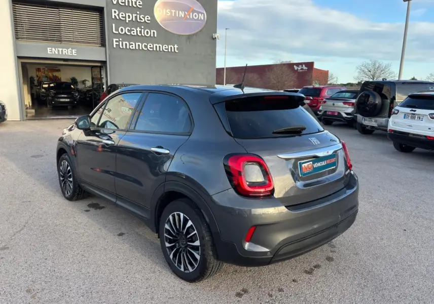 Vue 3/4 arrière droite d'une FIAT 500X gris métallisé avec jantes alliage et feux arrière LED, stationnée devant un concessionnaire.