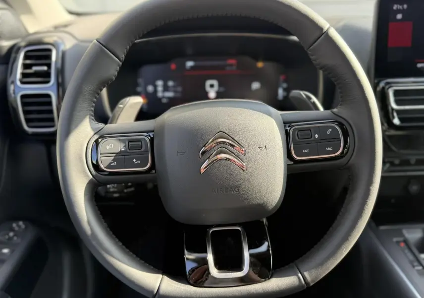 Vue rapprochée du volant noir en cuir du Citroën C5 Aircross 2025 avec commandes intégrées et tableau de bord numérique flou.