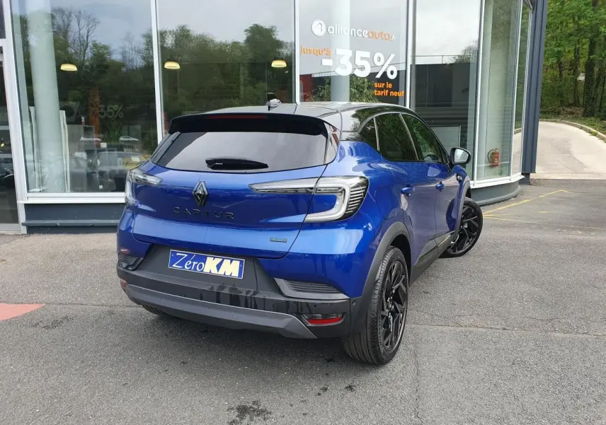 Vue 3/4 arrière droite d’un Renault Captur bleu Iron avec jantes noires et vitres surteintées, stationné devant un bâtiment.