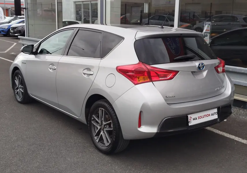 Vue 3/4 arrière droite d'une Toyota Auris HSD 136h gris aluminium, avec feux arrière distinctifs et vitres teintées.