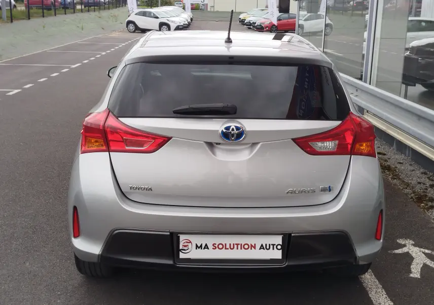 Vue arrière d'une Toyota Auris gris aluminium, mettant en valeur ses feux rouges et le logo hybride bleu.