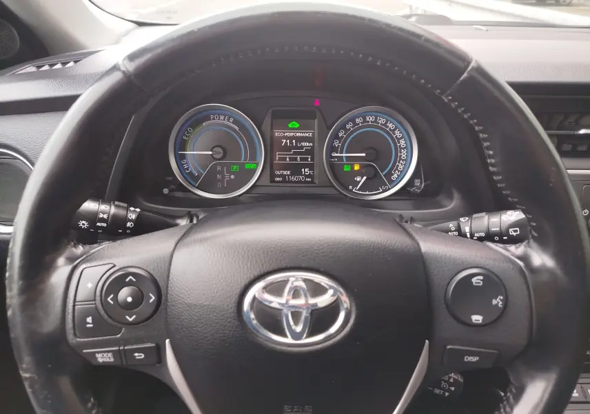 Vue intérieure centrée sur le volant noir de la Toyota Auris HSD 136h 2015 avec tableau de bord digital et compteurs bleus.
