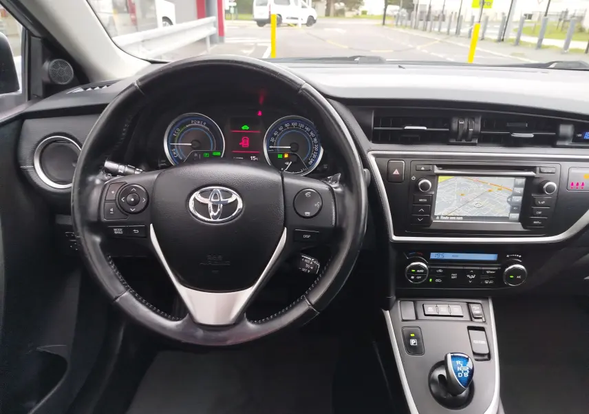 Vue intérieure du tableau de bord et volant noir de la Toyota Auris HSD 136h avec écran de navigation allumé.