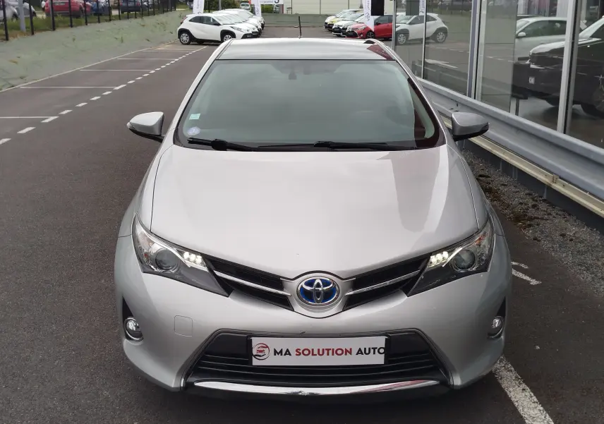 Vue avant d'une Toyota Auris HSD 136h gris aluminium avec phares allumés, stationnée devant une vitrine.