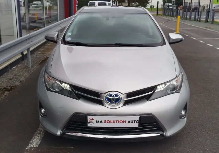 Vue avant d'une Toyota Auris HSD 136h gris aluminium, avec feux LED allumés et calandre noire brillante.
