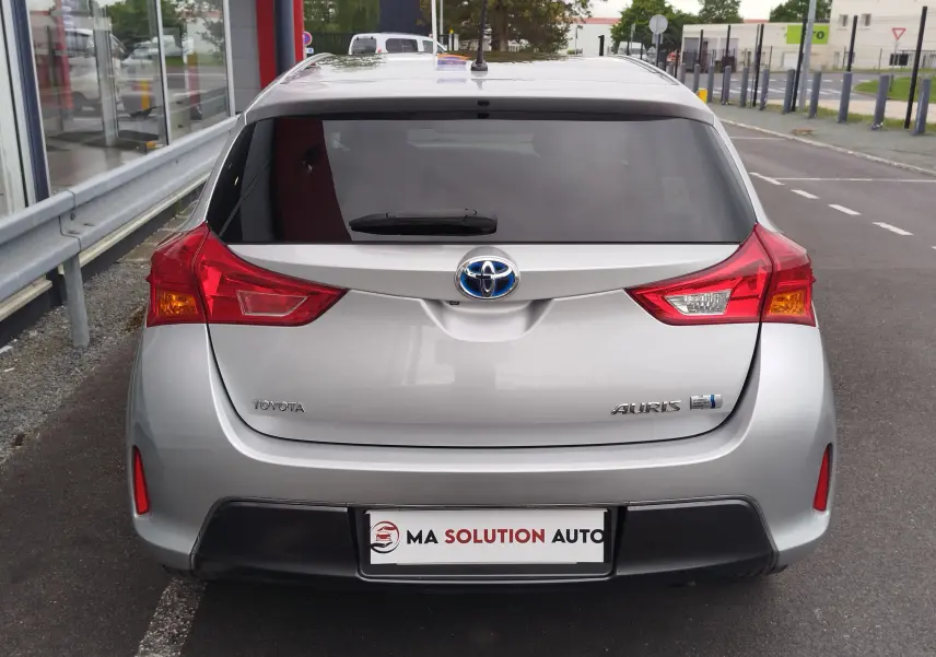 Vue arrière d'une Toyota Auris HSD 136h gris aluminium de 2015, avec feux arrière rouges et logo hybride visible.