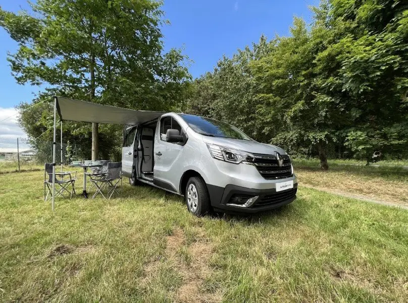 Vue 3/4 avant droit du Renault Trafic Fourgon gris avec porte latérale ouverte et auvent déployé sur herbe.