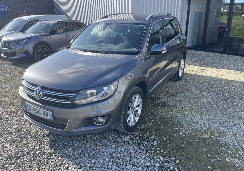 Volkswagen Tiguan gris foncé en 3/4 avant droit, avec jantes alliage et calandre chromée visible, stationné sur gravier.