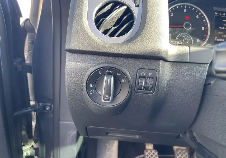 Détail du tableau de bord côté gauche du Volkswagen Tiguan gris foncé 2015, avec commande des phares et aérateur rond.