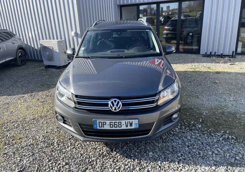 Vue avant d’un Volkswagen Tiguan gris foncé 2015, avec calandre chromée et phares allumés, stationné sur un sol gravillonné.