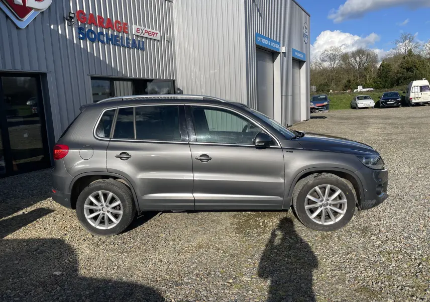 Profil droit d’un Volkswagen Tiguan gris foncé 2015 stationné devant un garage, avec jantes alliage visibles.