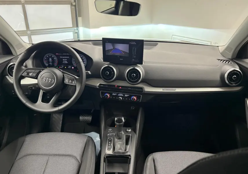 Vue intérieure avant de l'Audi Q2 35 TFSI S Line 2026, tableau de bord noir avec écran central et volant multifonction.