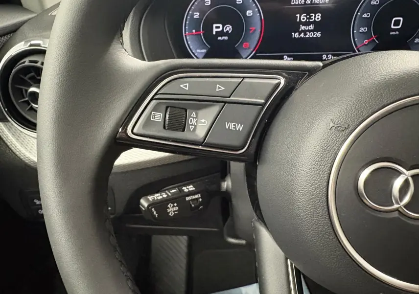 Gros plan sur le volant noir et le tableau de bord numérique de l'Audi Q2 35 TFSI S Line 2026, affichant l'heure et la vitesse.