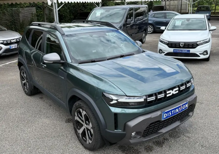 Vue 3/4 avant d’un Dacia Duster Hybrid 140 Journey vert cèdre avec calandre noire et jantes bicolores sur parking.