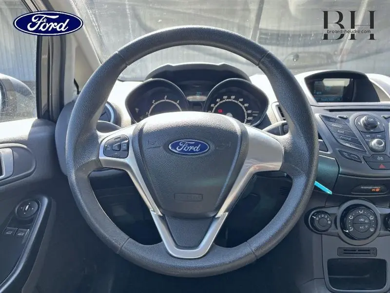 Vue intérieure centrée sur le volant de la Ford Fiesta noire 2015, avec tableau de bord et commandes visibles.