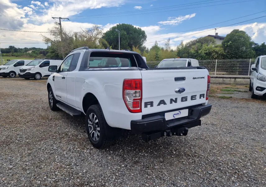 Ford Ranger Super Cabine blanc en 3/4 arrière droit, avec benne et logo Wildtrak visible sur un terrain gravillonné.