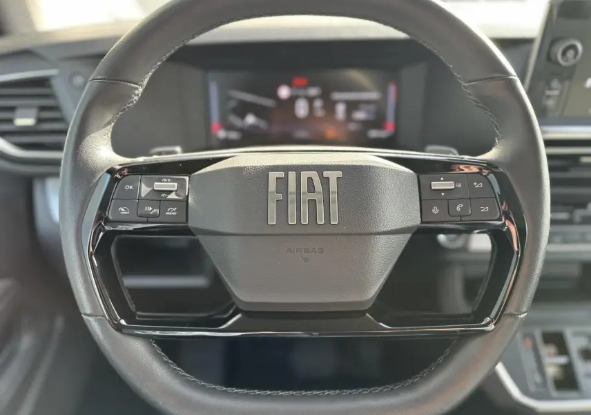 Vue rapprochée du volant noir FIAT avec commandes intégrées et tableau de bord numérique flou en arrière-plan.