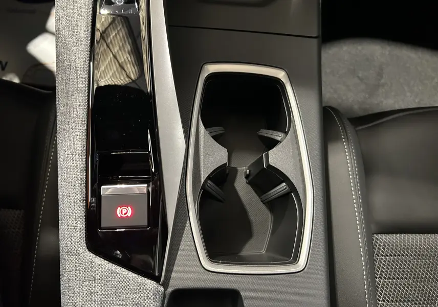 Vue plongeante sur la console centrale noire et grise du Peugeot 5008 III Hybrid avec porte-gobelets et frein à main électrique allumé.