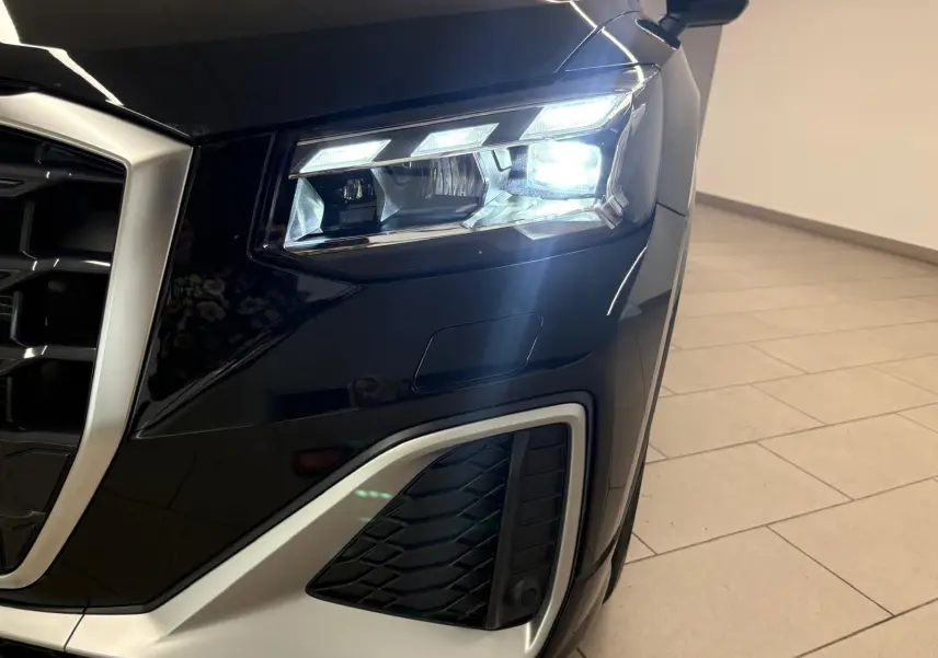 Gros plan sur le phare avant droit allumé de l'Audi Q2 35 TFSI S Line en Noir Mythic, avec calandre et détails argentés visibles.