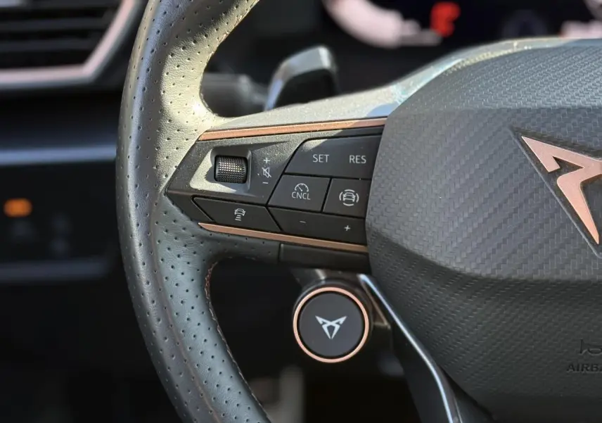 Gros plan sur le volant perforé noir du CUPRA LEON ST avec commandes intégrées et logo cuivre au centre.