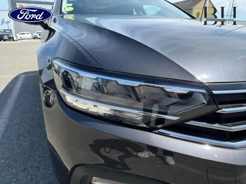 Gros plan sur le phare avant droit d'une Volkswagen Passat SW gris manganèse métallisé, avec calandre chromée visible.