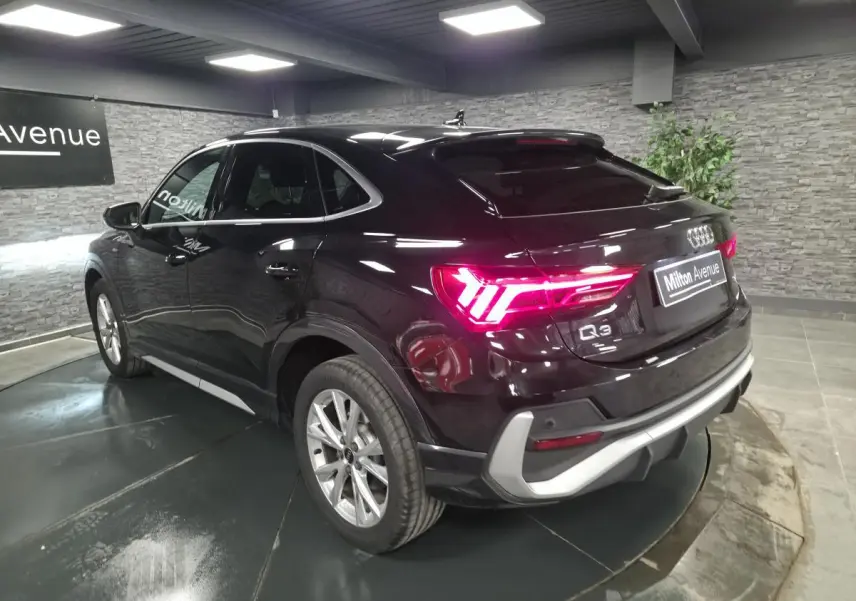 Audi Q3 Sportback noir vue 3/4 arrière droit, feux arrière allumés et vitres surteintées en intérieur showroom.