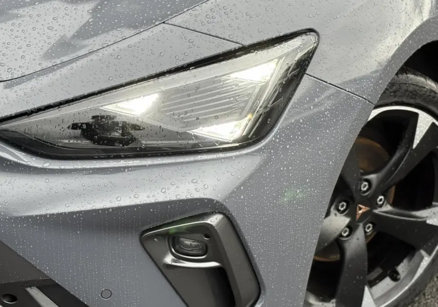 Gros plan sur le phare avant droit allumé et la jante noire de la CUPRA LEON ST gris Graphene sous la pluie.