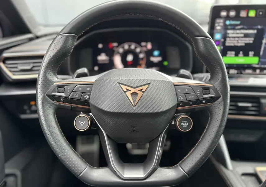 Volant cuir perforé noir avec logo CUPRA cuivré et commandes intégrées, tableau de bord numérique en arrière-plan.