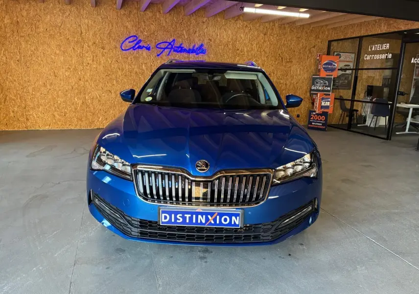 Vue de face d'une Skoda Superb Combi bleu métallisé, avec calandre chromée et phares LED dans un showroom.