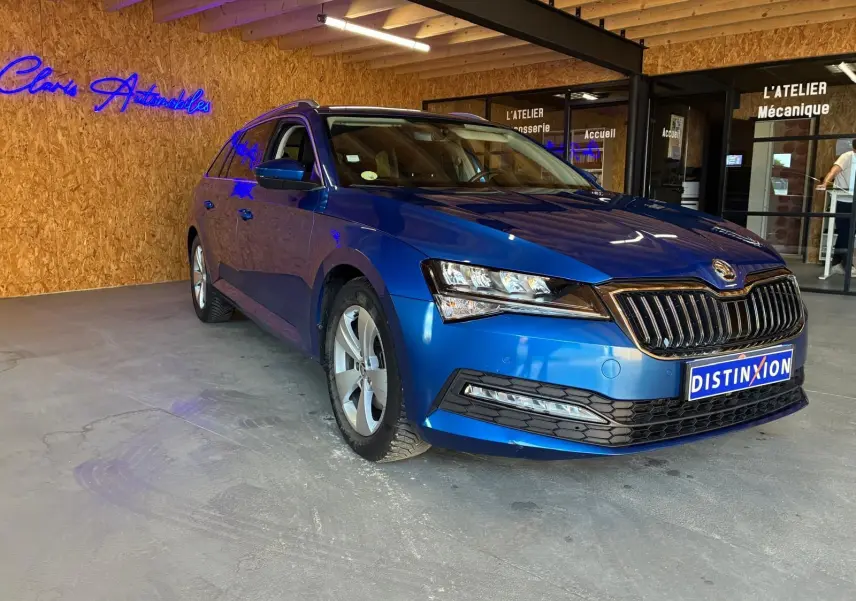 Skoda Superb Combi bleu en 3/4 avant droit, stationné en intérieur devant un mur en bois avec enseigne néon bleue.