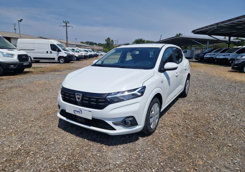 Dacia Sandero blanc vue 3/4 avant droit sur parking gravillonné, avec calandre noire et feux LED allumés.