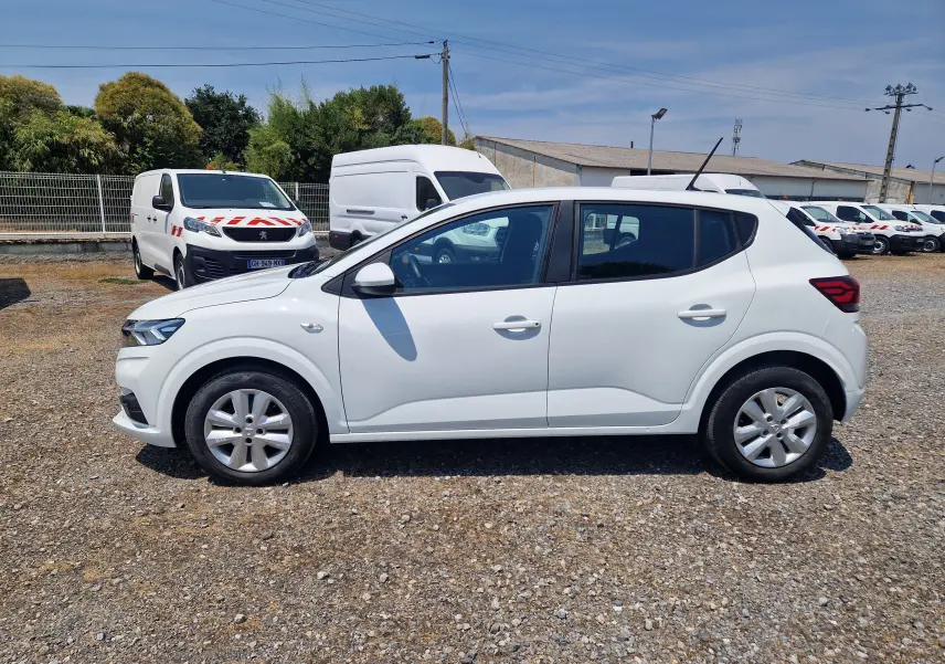 Vue latérale droite d'une Dacia Sandero blanche 2022, finition Confort, garée sur un sol gravillonné en extérieur.